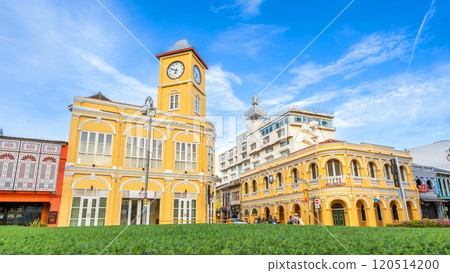 The Sino-Portuguese yellow clock tower and Chartered Bank building in Phuket Old Town, surrounded by lush greenery, stand out under a vivid blue sky, showcasing colonial charm 120514200