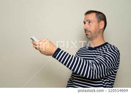 Bearded hispanic man in his 40s operating remote control, open air conditioner, saving energy. Isolated over beige background 120514604