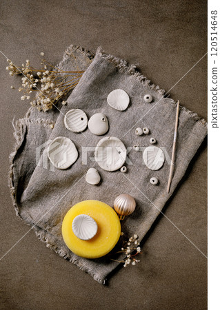 Ceramic workshop idea: handcrafted beads from clay or porcelain. Flat beads of raw white porcelain, tools on rough fabric. Small white flowers and jewelry findings. Dark background, top view. 120514648