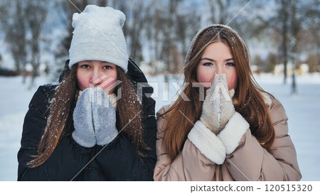 Two young girls in winter gear are breathing on their hands to keep warm in a snowy park 120515320