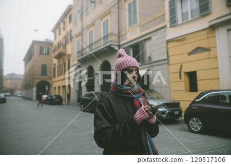 A woman in a pink hat and scarf stands on a street, looking at her phone 120517106
