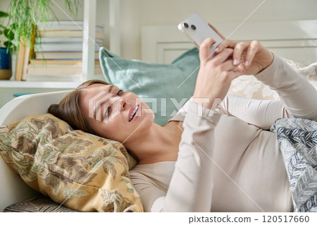Happy relaxed young woman lying on couch at home using smartphone 120517660