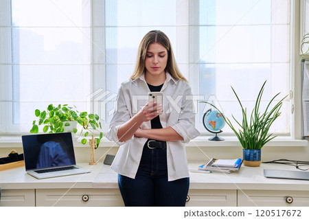 Young serious woman using smartphone standing at home near window 120517672