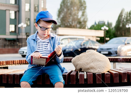 a 10-year-old boy wearing glasses and a cap sits on a bench on the street and reads an interesting book against the sky, copy space a 10-year-old boy wearing glasses and a cap sits on a bench on the street and reads an interesting book against the sky, copy space 120519700