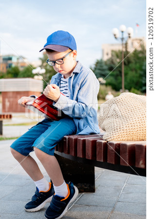 a 10-year-old boy wearing glasses and a cap sits on a bench on the street and reads an interesting book against the sky, copy space a 10-year-old boy wearing glasses and a cap sits on a bench on the street and reads an interesting book against the sky, copy space 120519702