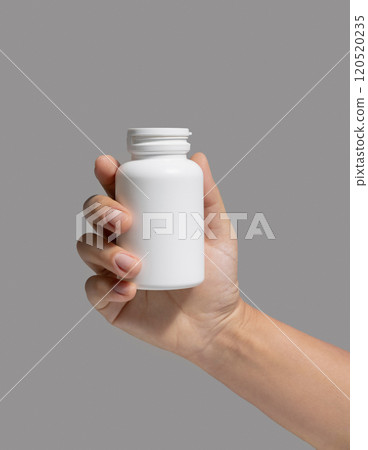 Female hand holding a white plastic pill bottle closeup against grey, Healthcare concept Female hand holding a white plastic pill bottle closeup against grey, Healthcare concept 120520235