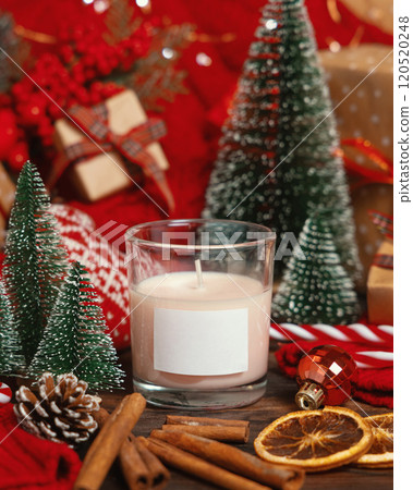 Glass candle with label near red knitted sweater and Christmas decor and lights, winter mockup 120520248
