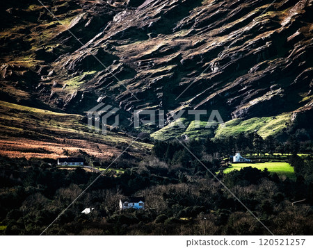 In a picturesque setting, small, white houses nestle among rolling green hills and rocky terrain during the tranquil evening hours, showcasing nature's beauty. 120521257