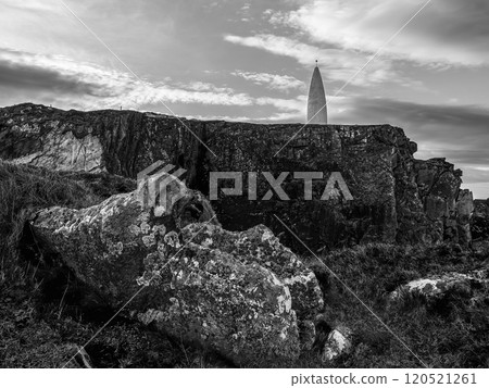 A rocky outcrop, partially vegetated, slopes upwards. A tall, white, conical structure is in the background. 120521261
