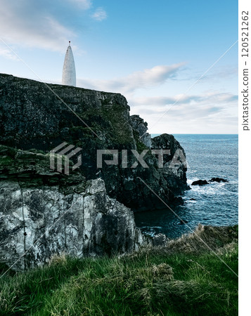 A lighthouse stands on a rocky cliff overlooking the sea, surrounded by grass. The sky is partly cloudy, adding a serene atmosphere to the coastal setting. 120521262