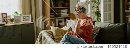 Elderly woman sitting on sofa, talking on phone with smile on face Shelves with books and photo frames visible in background, creating warm, homey environment 120521418