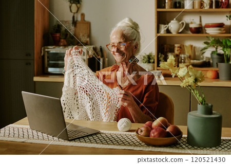Senior woman smiling and displaying a piece of knitted fabric through laptop, sitting in cozy kitchen with various plants and fruit bowl. Wooden utensils and ceramics decorating kitchen space 120521430