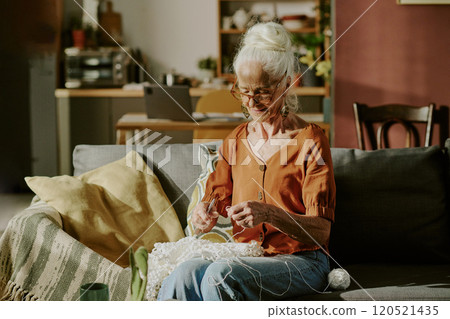 Senior woman engaged in knitting while sitting on a comfortable couch in well-lit, cozy living room featuring home decor elements and warm atmosphere Senior woman engaged in knitting while sitting on a comfortable couch in well-lit, cozy living room featuring home decor elements and warm atmosphere 120521435
