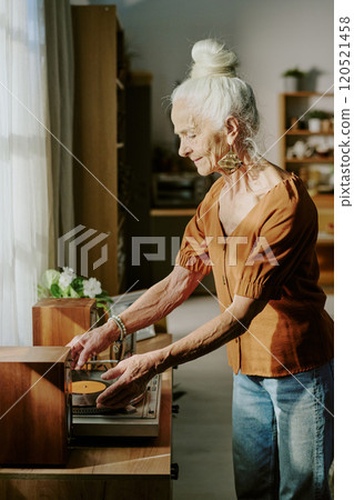 Elderly woman listening to music on a vintage record player during sunny day in cozy room with wooden furniture and plants, looking content and relaxed while turning knob Elderly woman listening to music on a vintage record player during sunny day in cozy room with wooden furniture and plants, looking content and relaxed while turning knob 120521458