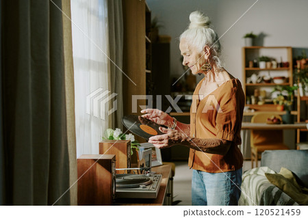 Senior woman standing in living room, adjusting record player. Room is decorated with plants and bookshelves, creating a peaceful atmosphere. Sunlight streaming through windows 120521459