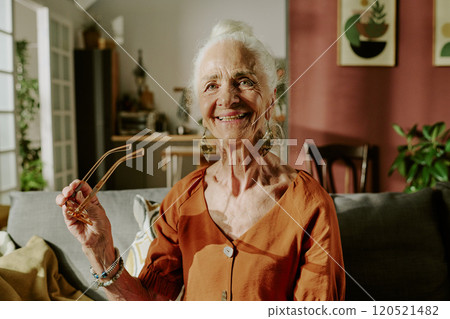 Elderly woman standing in living room, smiling while holding glasses. Cozy interior features plants, artwork, and modern furniture creating homey atmosphere 120521482