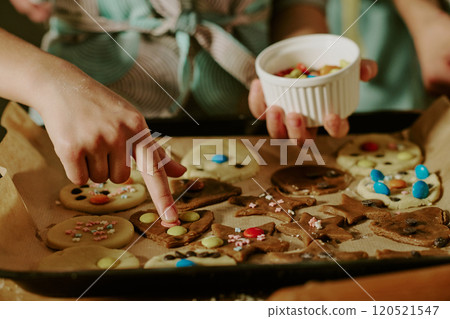 Person decorating a variety of holiday cookies on baking sheet wearing apron focusing on individual cookie designs holding cup of toppings 120521547