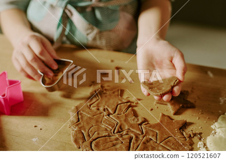 Hands cutting gingerbread dough on table, using star-shaped cookie cutters, creating festive shapes. Dough and flour are spread on surface, with some cookie shapes already cut and lifted Hands cutting gingerbread dough on table, using star-shaped cookie cutters, creating festive shapes. Dough and flour are spread on surface, with some cookie shapes already cut and lifted 120521607