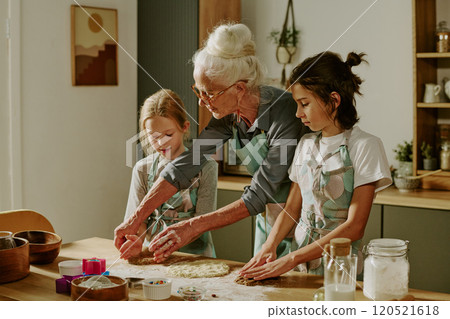 Senior woman guiding two young girls in kitchen while preparing dough together, focused on baking cookies and enjoying the moment without distractions 120521618