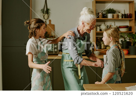 Elderly woman teaching two young girls how to bake while smiling and wearing aprons in cozy kitchen setting with various cooking utensils and ingredients around Elderly woman teaching two young girls how to bake while smiling and wearing aprons in cozy kitchen setting with various cooking utensils and ingredients around 120521620