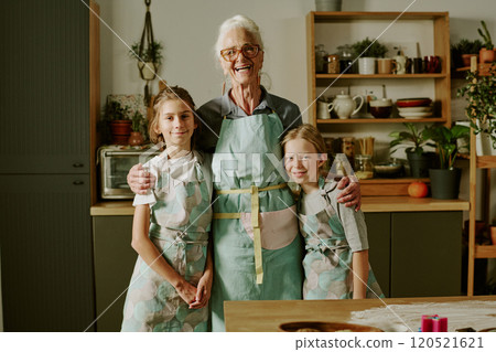 Senior woman baking with two young girls, wearing aprons and smiling together in kitchen, standing near wooden table with baking supplies Senior woman baking with two young girls, wearing aprons and smiling together in kitchen, standing near wooden table with baking supplies 120521621