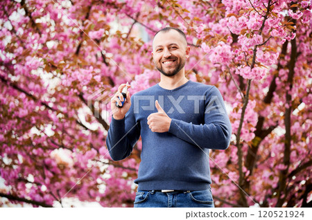 Man allergic using medical nasal drops, suffering from seasonal allergy at spring in blossoming garden. Handsome smiling man showing thumbs up near blooming tree outdoors. Spring allergy concept. Man allergic using medical nasal drops, suffering from seasonal allergy at spring in blossoming garden. Handsome smiling man showing thumbs up near blooming tree outdoors. Spring allergy concept. 120521924