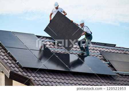 Workers building solar panel system on roof of house. Two men installers in helmets carrying photovoltaic solar module outdoors. Alternative, green and renewable energy generation concept. Workers building solar panel system on roof of house. Two men installers in helmets carrying photovoltaic solar module outdoors. Alternative, green and renewable energy generation concept. 120521953