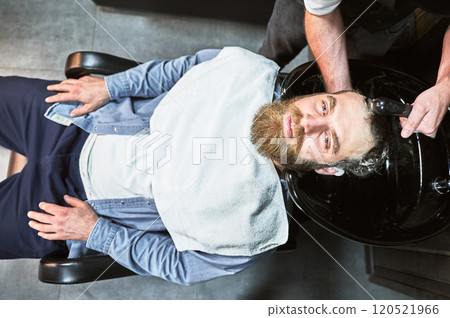 Ginger-haired man getting his hair washed. Male hairdresser washing hair with deep cleaning products. Handsome client relaxing with towel on chest. 120521966