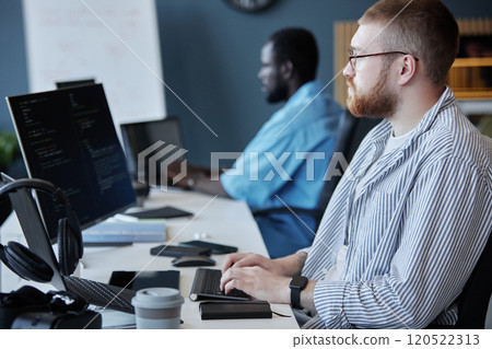 Multi-ethnic group of developers focused on coding and discussing project details in a modern office setting. Senior programmer working on computer screen with tech gadgets nearby 120522313