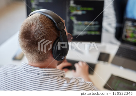 Person sitting at desk and wearing black over-ear headphones while focusing on coding on computer screen with code in background 120522345