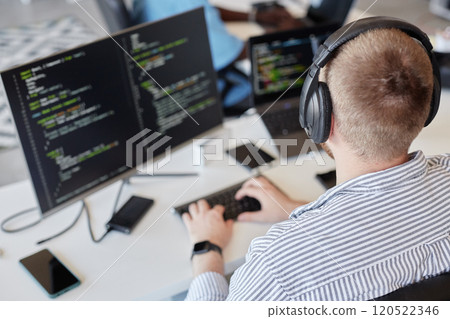 Man wearing headphones sitting at desk working on coding project on dual monitors. Image shows work environment with various devices and tools surrounding him Man wearing headphones sitting at desk working on coding project on dual monitors. Image shows work environment with various devices and tools surrounding him 120522346