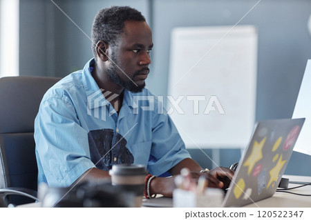 Man concentrating while working on modern laptop in office setting, surrounded by various office supplies and equipment, a whiteboard in background 120522347