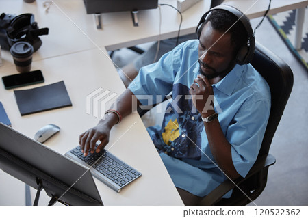 African American man focusing on computer work, wearing headphones, seated at desk in modern office environment with various office supplies placed around. 120522362