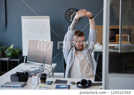 Employee seated at desk stretching arms in a modern office surrounded by technology, such as computer screens and VR headsets having whiteboard and clock on wall background 120522368