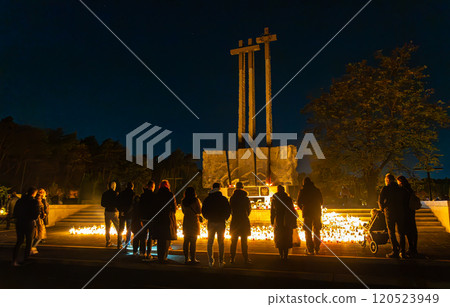 A solemn night scene of people gathered around a monument, lighting candles to honor the departed during All Saints Day. The warm candlelight contrasts with the dark sky  120523949
