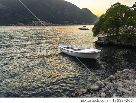 A small white boat gently floating on calm waters by a rocky shore, framed by trees and mountains under the glow of sunset. A small white boat gently floating on calm waters by a rocky shore, framed by trees and mountains under the glow of sunset. 120524205