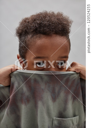 Vertical shot of funny little Black boy hiding face in t shirt pulling up collar and peeking out at camera, while standing against gray background in studio Vertical shot of funny little Black boy hiding face in t shirt pulling up collar and peeking out at camera, while standing against gray background in studio 120524255