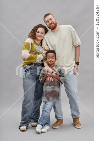 Vertical full length shot of happy adoptive parents posing in gentle hug with little Black boy while standing against gray background in studio 120524267