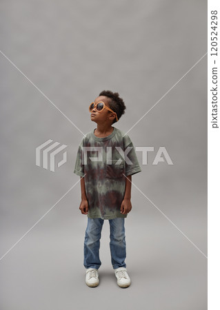 Vertical shot of little Black boy in casual outfit and stylish sunglasses looking away while standing against gray background in studio, copy space Vertical shot of little Black boy in casual outfit and stylish sunglasses looking away while standing against gray background in studio, copy space 120524298
