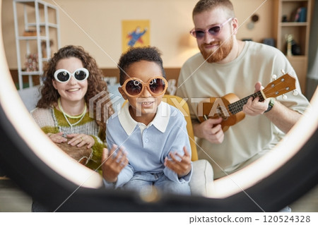 Smiling little Black boy in sunglasses shooting video with creative parents for family channel in social network playing music as band, while looking at camera through ring light lamp at home Smiling little Black boy in sunglasses shooting video with creative parents for family channel in social network playing music as band, while looking at camera through ring light lamp at home 120524328