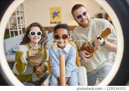 Cheerful little Black boy in sunglasses shooting video with creative parents for family channel in social network playing music as band, while looking at camera through ring light lamp at home Cheerful little Black boy in sunglasses shooting video with creative parents for family channel in social network playing music as band, while looking at camera through ring light lamp at home 120524331