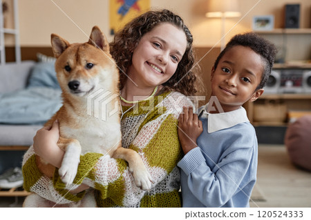Portrait of cheerful young woman posing together with little Black boy while holding cute dog in bright room at home, happy adoptive family concept 120524333