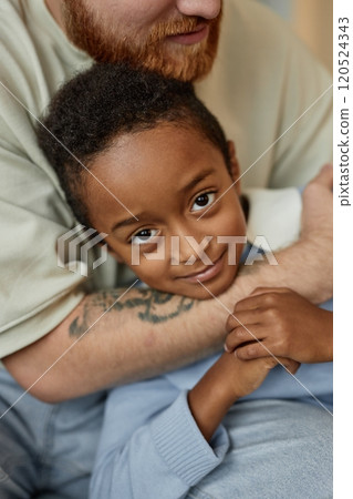 Vertical portrait shot of little smiling Black boy in tender embrace of adoptive father happily looking at camera while spending time with parent at home 120524343