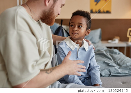 Medium shot of little Black boy listening with attention to supportive adoptive father during important conversation while sitting on bed in kids room, copy space 120524345