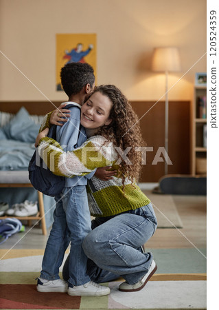 Vertical shot of smiling adoptive mother giving gentle hug to Black boy in bedroom raising happy confident kid while sending son off to school, copy space 120524359