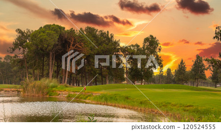 A serene pond reflecting the vibrant sunset sky, surrounded by lush greenery, trees, and a peaceful golf course. 120524555