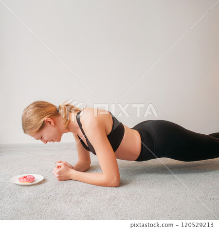 High motivation. Sport plank. Young girl in black suit doing elbow exercise for core strengthening on fitness mat looking at plate with cake in modern living room interior. 120529213