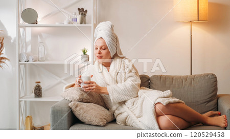Salon procedure. Spa weekend. Inspired young woman in bath towel white soft bathrobe with coffee mug enjoying rest on couch at home after shower. Salon procedure. Spa weekend. Inspired young woman in bath towel white soft bathrobe with coffee mug enjoying rest on couch at home after shower. 120529458