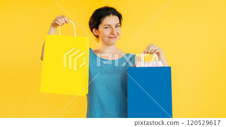 Happy woman in blue shirt happily holding yellow blue shopping bags isolated on vibrant yellow color empty space background. Happy woman in blue shirt happily holding yellow blue shopping bags isolated on vibrant yellow color empty space background. 120529617