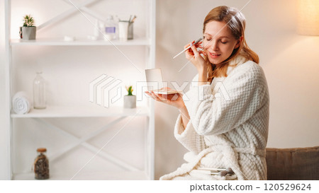 Cosmetic morning. Beauty routine. Happy woman applying makeup using powder brush in white robe enjoying process. Cosmetic morning. Beauty routine. Happy woman applying makeup using powder brush in white robe enjoying process. 120529624